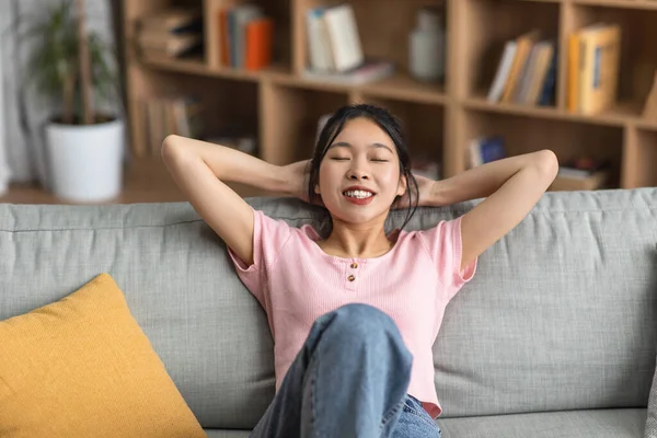 Life is good. Carefree asian lady leaning back on sofa with closed eyes and hands behind head, resting at home — Foto de Stock