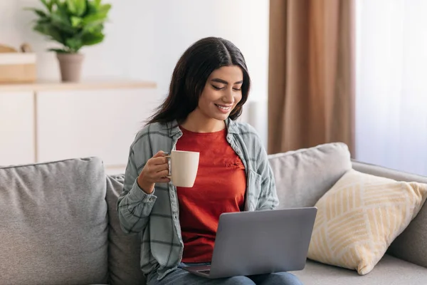 Happy Arab businesswoman having online business meeting on laptop, drinking coffee at home office — Stock fotografie