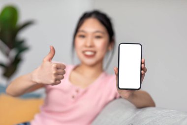 Genç Asyalı bayan evdeki kanepede oturuyor, beyaz beyaz ekranlı akıllı telefonu gösteriyor ve başparmağını kaldırıyor.