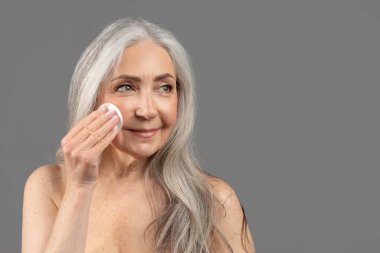 Gri stüdyo arkaplanı ve boş alan üzerindeki makyajı temizlemek için pamuk pedi kullanan sevimli yaşlı kadın.