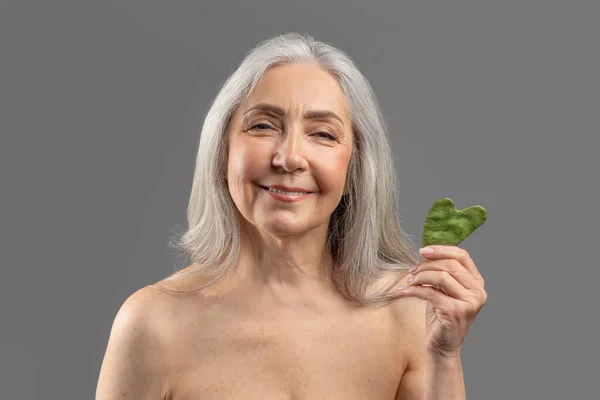 Lovely senior Caucasian woman holding gua sha lymphatic drainage facial self massager and smiling at camera — 스톡 사진