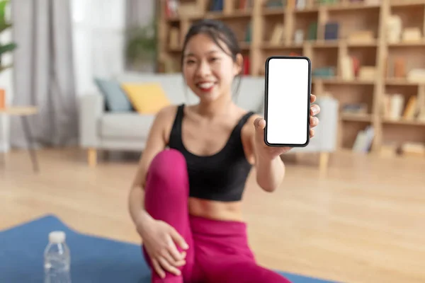 Glücklich athletisch koreanische Dame in Sportbekleidung sitzt auf Yogamatte zu Hause und zeigt modernes Handy mit leerem Bildschirm — Stockfoto