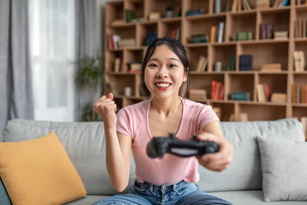 快乐的韩国女士，带着控制器玩网络游戏，做"是"的手势，坐在家里的沙发上 — 图库照片