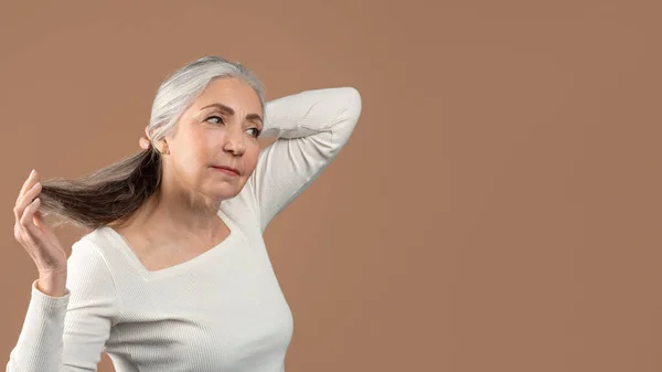Beautiful mature lady touching her long grey hair and looking aside at empty space on brown studio background, banner — Foto Stock