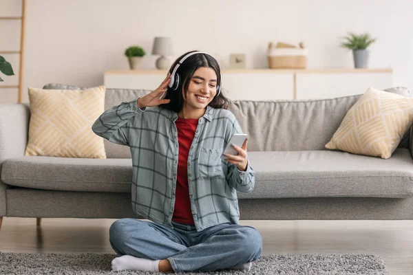 Ritratto di allegra giovane araba in cuffia che ascolta musica su dispositivo mobile, seduta sul pavimento di casa — Foto Stock