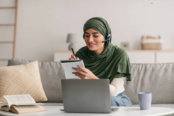 在家中用笔记本电脑、戴耳机、在远程课堂上写下信息的快乐的阿拉伯妇女 — 图库照片