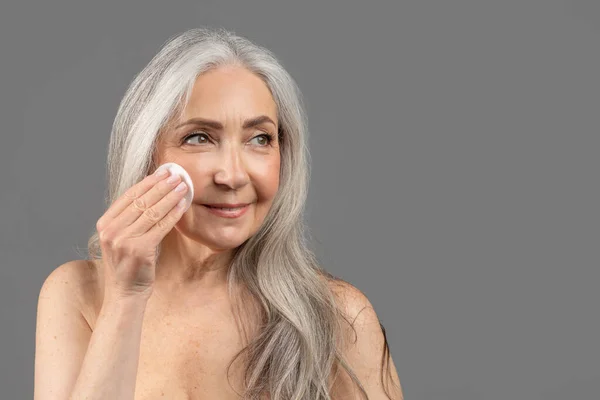 Lovely senior woman using cotton pad to remove makeup over grey studio background, empty space — стоковое фото