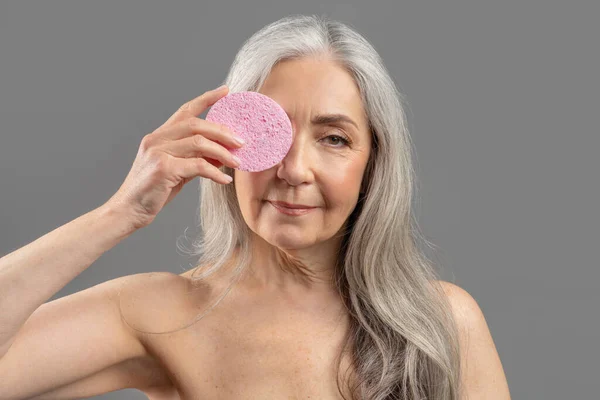 Lovely senior lady holding cosmetic sponge in front of her eye, ready for face cleansing or removing makeup —  Fotos de Stock