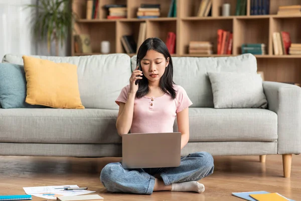 Asian female freelancer talking on cellphone and typing on laptop, working online from home, sitting on floor — Foto de Stock