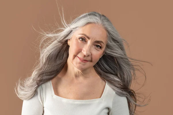 Retrato de mulher sênior sorridente posando com cabelos longos e saudáveis cinza soprando voando no fundo do estúdio marrom — Fotografia de Stock