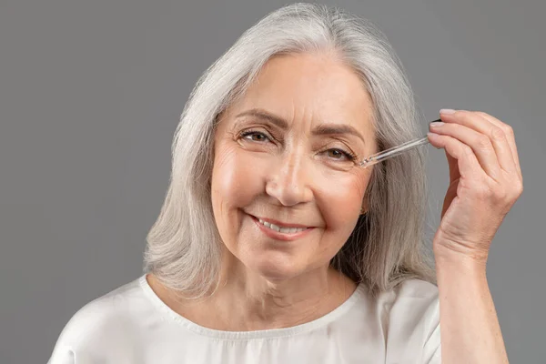 Rejuvenescer cosméticos. Mulher sênior bonita aplicando soro anti-envelhecimento em seu rosto sobre fundo estúdio cinza — Fotografia de Stock