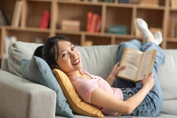 Весела азіатська леді відпочиває на дивані, читає книгу, проводячи час вдома у вихідні, посміхаючись на камеру — стокове фото