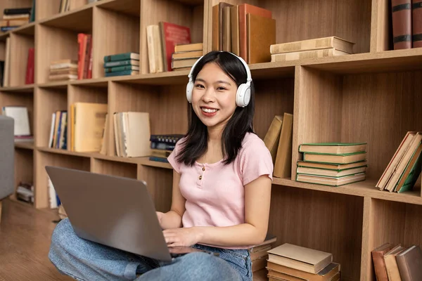 Happy asian lady in wireless headphones typing on laptop keyboard while leaning on bookshelf and smiling at camera — Stockfoto