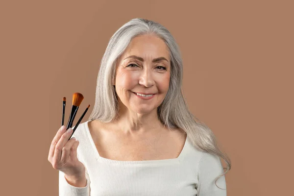 Retrato de bela mulher madura segurando conjunto de diferentes escovas cosméticas sobre fundo de estúdio marrom — Fotografia de Stock