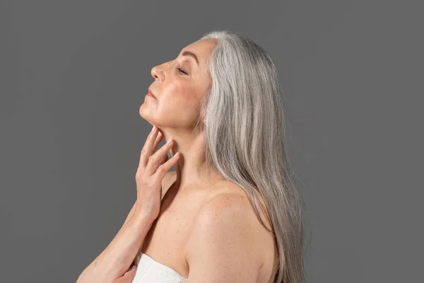 Feminine senior lady with silky skin wearing towel, touching her neck on grey studio background, side view portrait — 스톡 사진