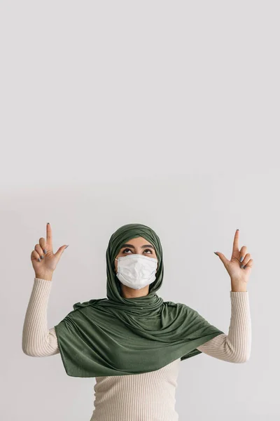 Coronavirus protection. Young Arab woman in hijab and antiviral face mask pointing up at empty space over her head — Foto Stock