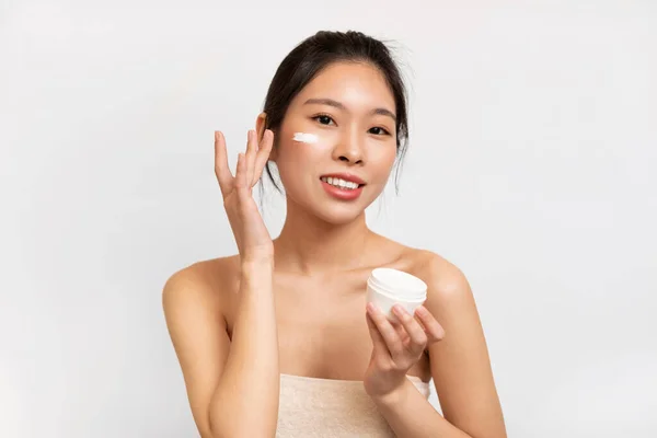 Portrait of Asian lady applying cream on cheek holding jar — Stok fotoğraf