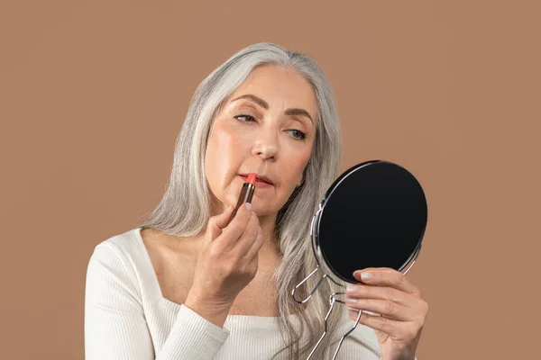 Lovely mature lady applying lipstick in front of mirror, taking care of herself on brown studio background — Stock Photo, Image