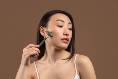 Asyalı genç bayan yeşil taştan bir yüz silindiri kullanıyor. Kahverengi stüdyo arka planında poz verirken yana bakıyor.