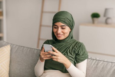 tesettürde oturan tesettürlü genç bir Arap kadın cep telefonuyla sosyal ağları kontrol ediyor, evden internete bakıyor.