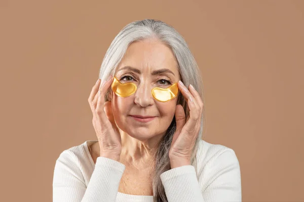 Kahverengi stüdyo arka planında kozmetik göz bandı takan yaşlı kadın. — Stok fotoğraf