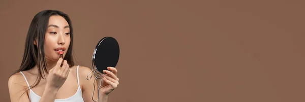 Pretty asian lady making daily makeup, using mirror and lipstick over brown studio background, panorama with free space — Stock Photo, Image