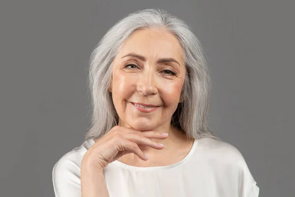Yaşlı bir kadının güzel portresi yüzüne dokunuyor, kameraya bakıyor ve gri stüdyo arka planında gülümsüyor. — Stok fotoğraf
