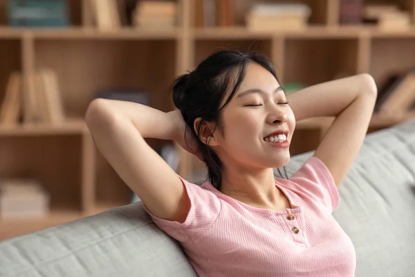 Rest and relax in your own home. Happy asian lady with closed eyes threw hands behind head sitting on sofa — ストック写真