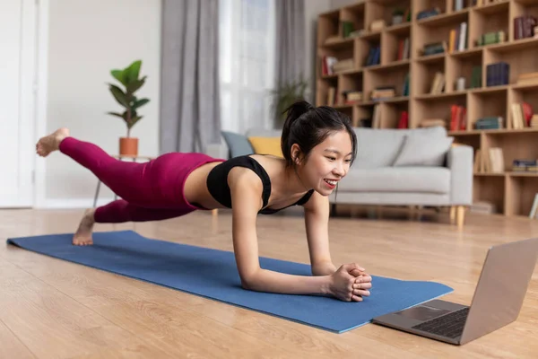 Fit asian woman standing in elbow plank, lifting her leg, exercising at home and following online video tutorial — Stockfoto