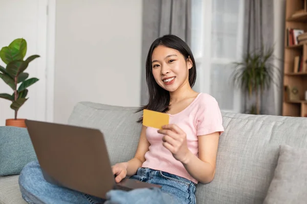 Excited asian lady sitting on sofa, using laptop and holding credit card, shopping online from home, copy space — Foto Stock