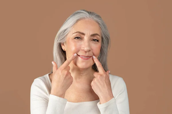 Gyönyörű idős nő megható sarkaiban mosoly ujjaival barna stúdió háttér — Stock Fotó