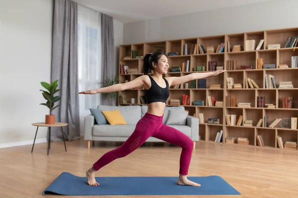 Concepto de yoga doméstico. Slim joven asiática dama haciendo embestida, de pie en el guerrero asana mientras que el entrenamiento en casa, longitud completa — Foto de Stock