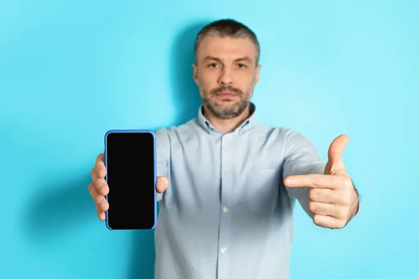 Serious Male Showing Cellphone Screen Pointing Finger Over Blue Background — стоковое фото