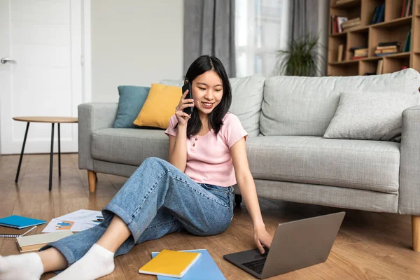 Senhora coreana feliz falando no smartphone e olhando para a tela do laptop, sentado no chão perto do sofá em casa, espaço de cópia — Fotografia de Stock