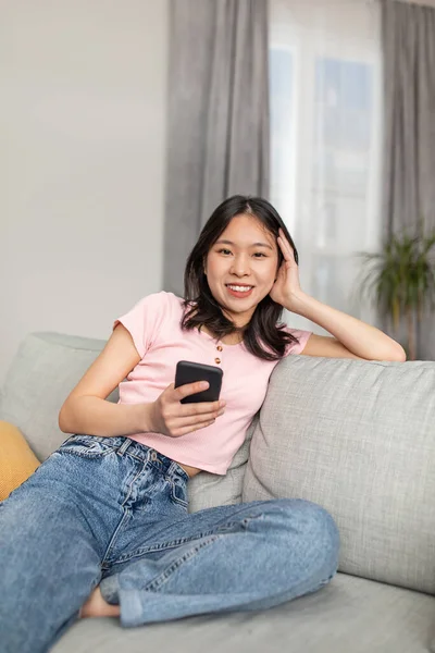 Happy asian lady sitting on sofa in living room and using smartphone, checking social networks, resting at home — ストック写真