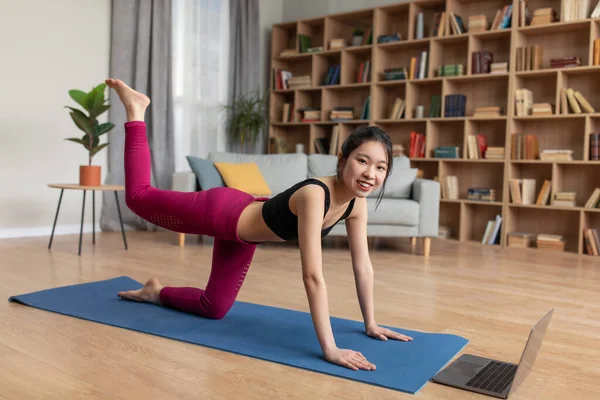 Çevrimiçi ev içi antrenman. Kalça ve bacak egzersizi yapan mutlu Asyalı kadın dizüstü bilgisayarın yanında dört ayak üstünde egzersiz yapıyor.
