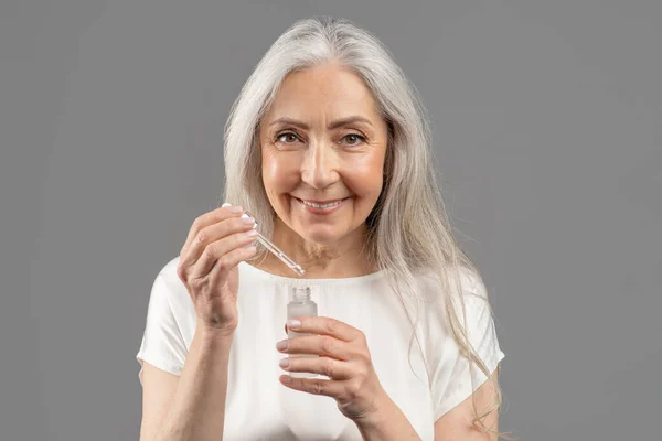 Догляд за шкірою та здоров'я. Портрет усміхненої старшої жінки, що застосовує сироватку обличчя з крапельницею на сірому студійному фоні — стокове фото