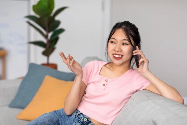 Excited asian lady speaking on cellphone while resting on couch, having conversation on mobile device — Foto de Stock