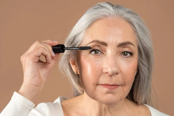 Beauty portrait of charming senior woman applying mascara on her lashes over brown studio background — Stock Photo, Image