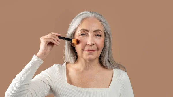 Natural makeup for mature skin. Charming senior lady putting on organic cosmetics with brush on brown studio background — Foto de Stock