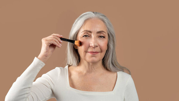 Natural makeup for mature skin. Charming senior lady putting on organic cosmetics with brush on brown studio background