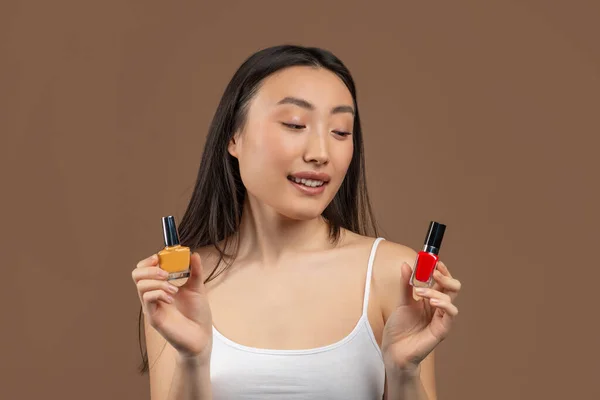 Portrait of young asian woman hesitating about choosing nail polish, posing over brown studio background — Stock Photo, Image