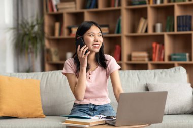 Pozitif Koreli kadın evdeki koltukta otururken telefonla konuşuyor ve dizüstü bilgisayar kullanıyor.