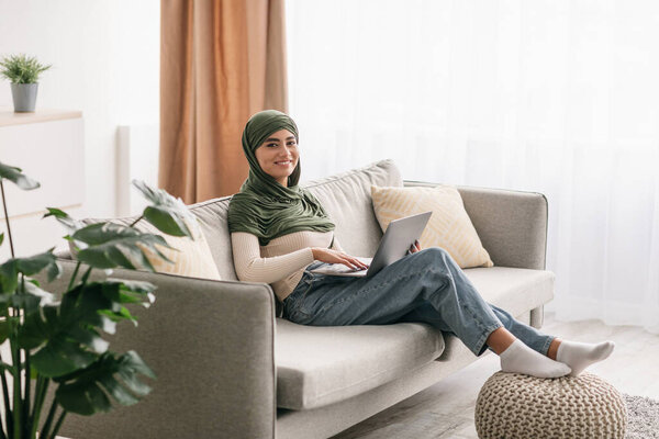 Full length of joyful young Arab woman in hijab working on laptop, sitting on couch at home office, copy space