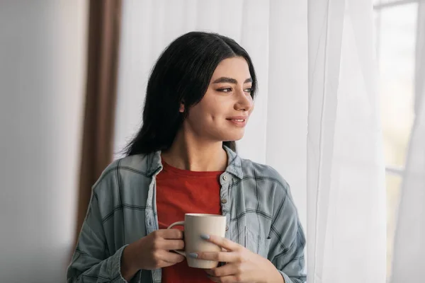 Positive young Arab woman drinking hot coffee, looking out window at home, enjoying peaceful morning — Stockfoto