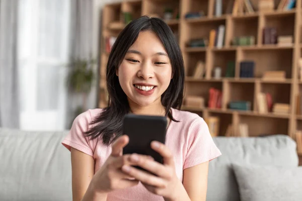 Positive koreanische Dame mit Smartphone, Surfen im Internet, Chat online, Video ansehen, auf der Couch zu Hause sitzen — Stockfoto