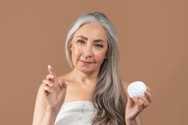 Lovely senior woman applying moisturizing cream under her eyes on brown studio background — Stock Photo, Image