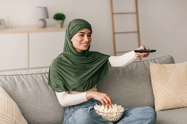 Happy young Arab woman with remote control sitting on couch, watching TV and eating popcorn at home — Stok Foto