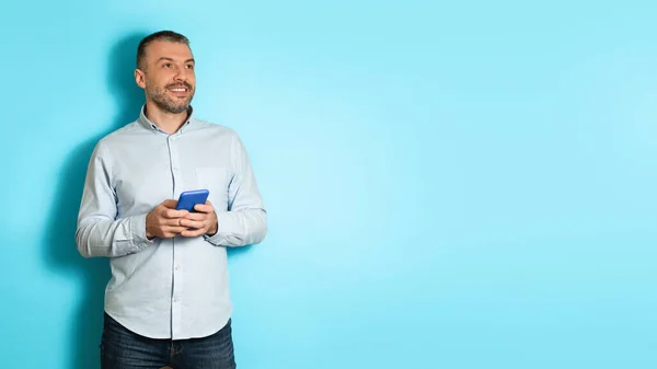 Mann mittleren Alters hält Handy in der Hand und blickt über blauen Hintergrund hinweg — Stockfoto