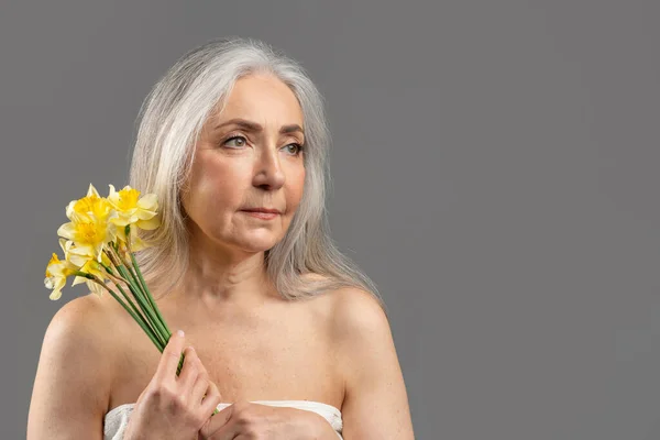 Портрет чарівної старшої леді, що тримає нарциси і дивиться на порожній простір на сірому студійному фоні — стокове фото